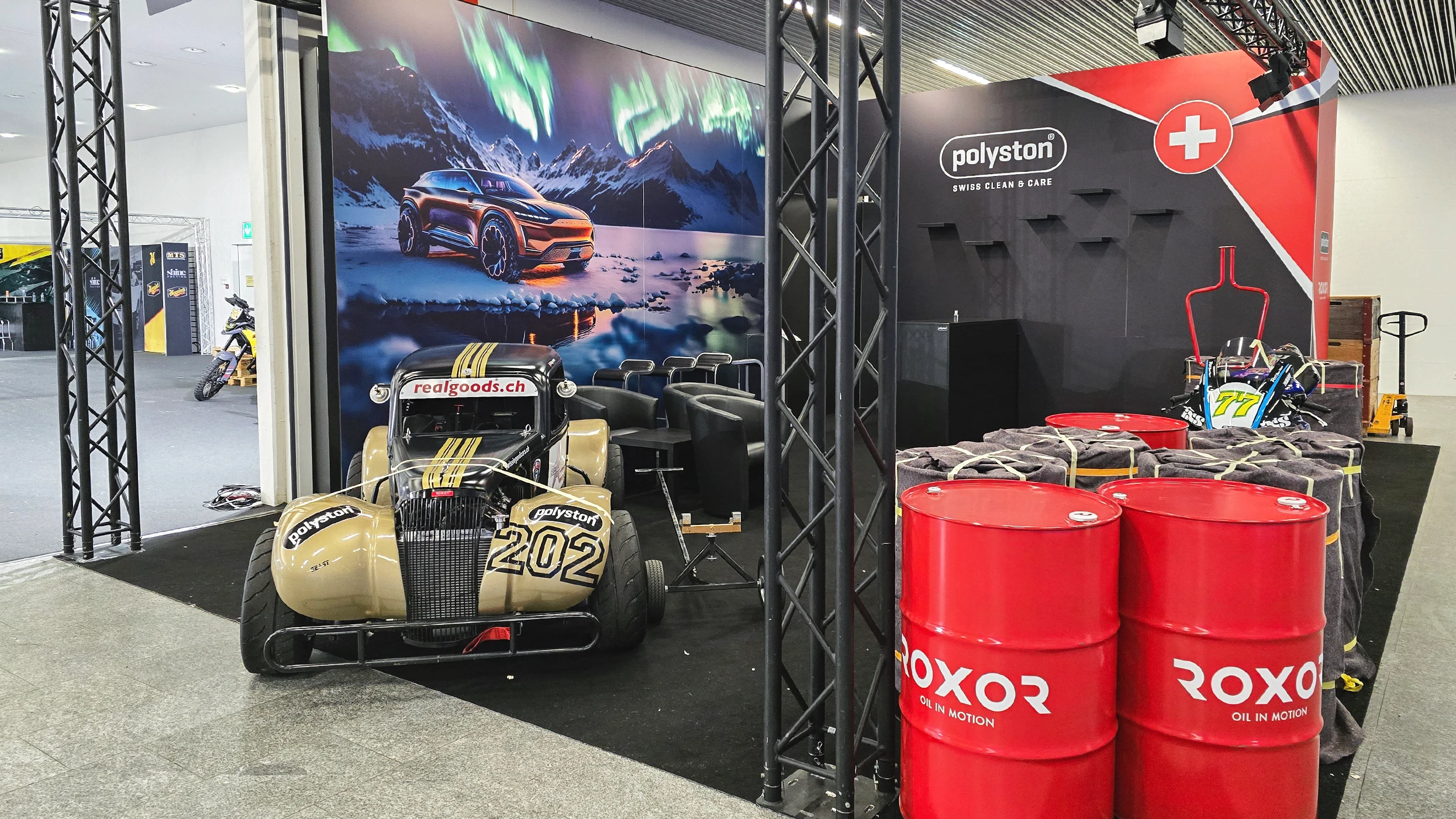 Exhibition stand with a classic black-and-beige racing car, No. 202, at the front. A large mural shows an SUV in a polar landscape with the Northern Lights. Right: two red oil drums and go-kart No. 77 by a black Polyston stand.