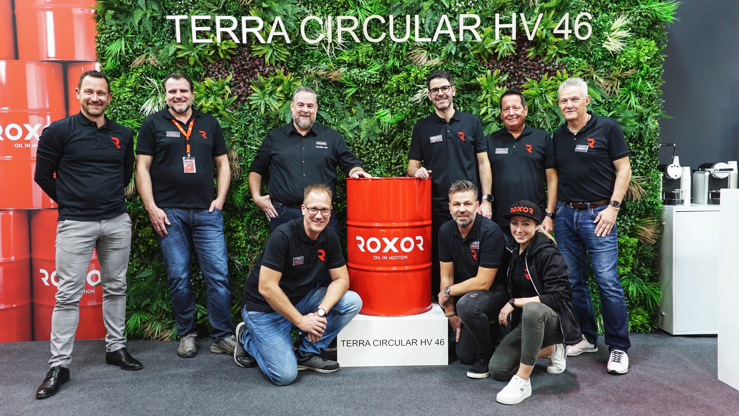 Nine smiling people (eight men, one woman) are standing in front of a green wall bearing the words ‘TERRA CIRCULAR HV 46’. In the centre stands a red ROXOR oil drum on a plinth bearing the same text. They are all wearing black company T-shirts with logos.
