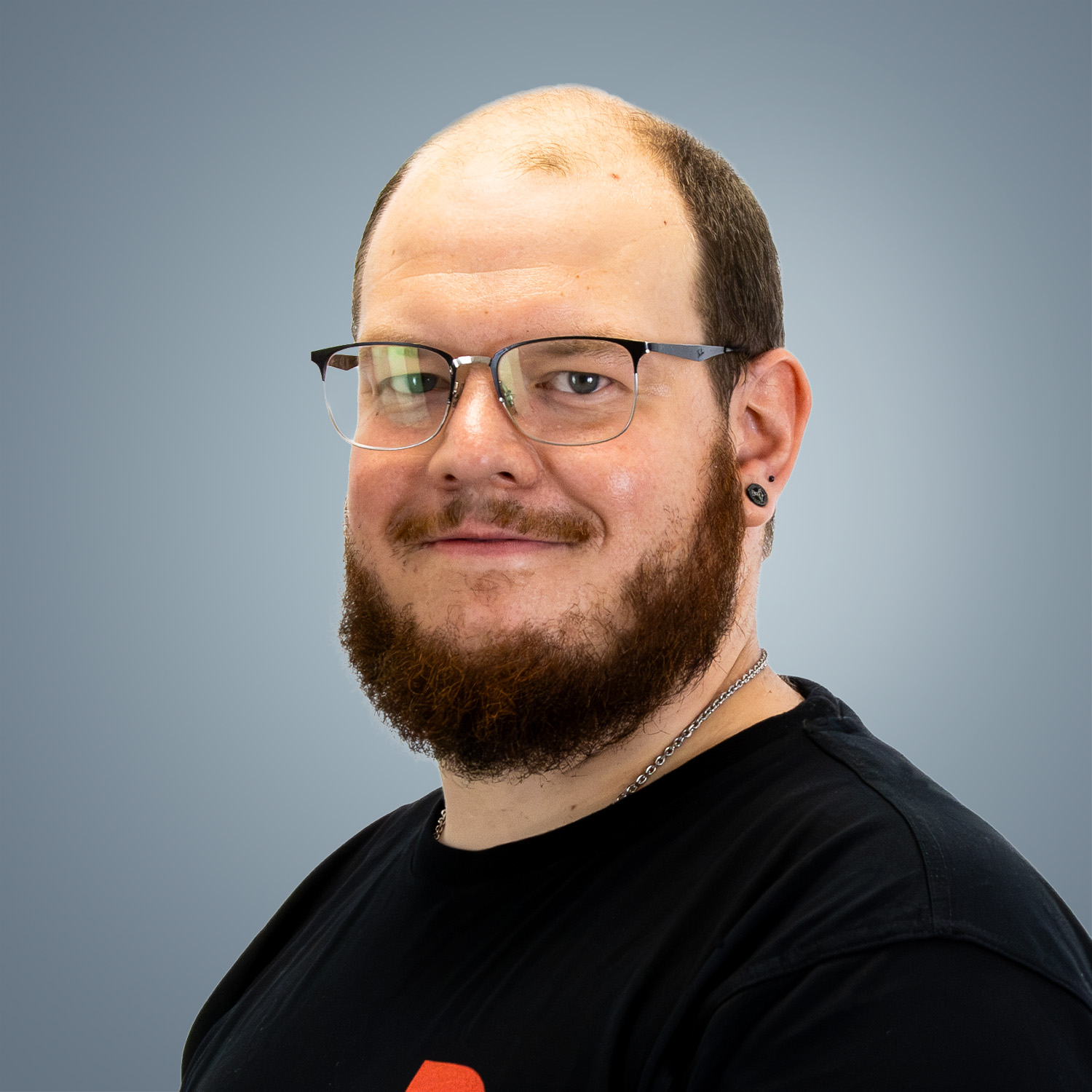 A smiling man with a reddish-brown full beard and a bald head is wearing black glasses and has a black ear piercing. He is looking straight at the camera and is wearing a black T-shirt and a silver necklace. There is a grey gradient in the background.