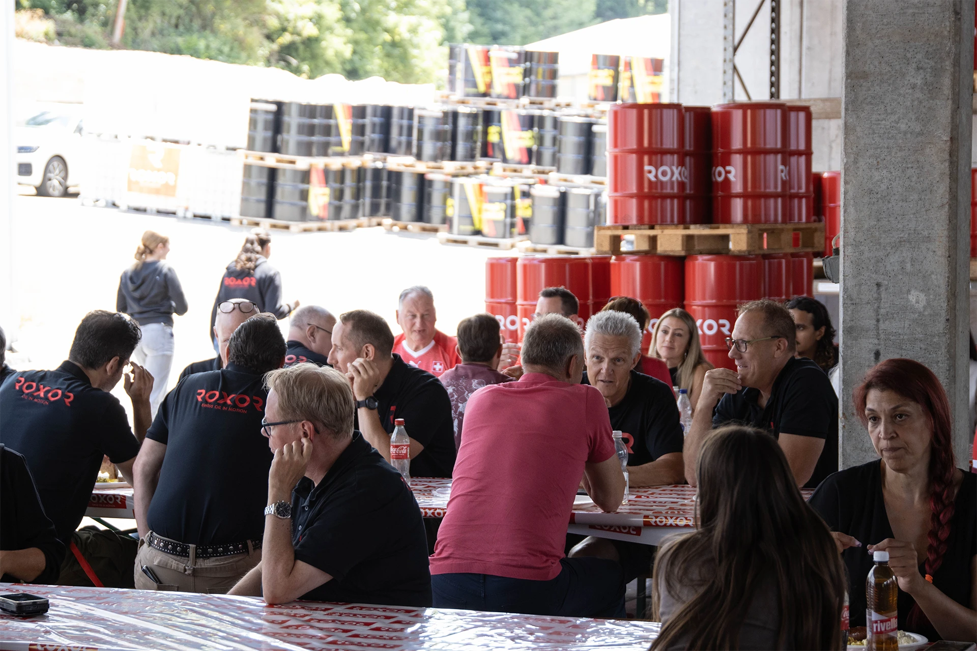 Erwachsene sitzen an Tischen und unterhalten sich. Viele tragen schwarze T-Shirts mit „ROXOR“-Logo. Im Hintergrund sind hohe Stapel roter und schwarzer Industriefässer mit „ROXOR“-Schriftzug zu sehen, was auf ein Firmentreffen hindeutet.