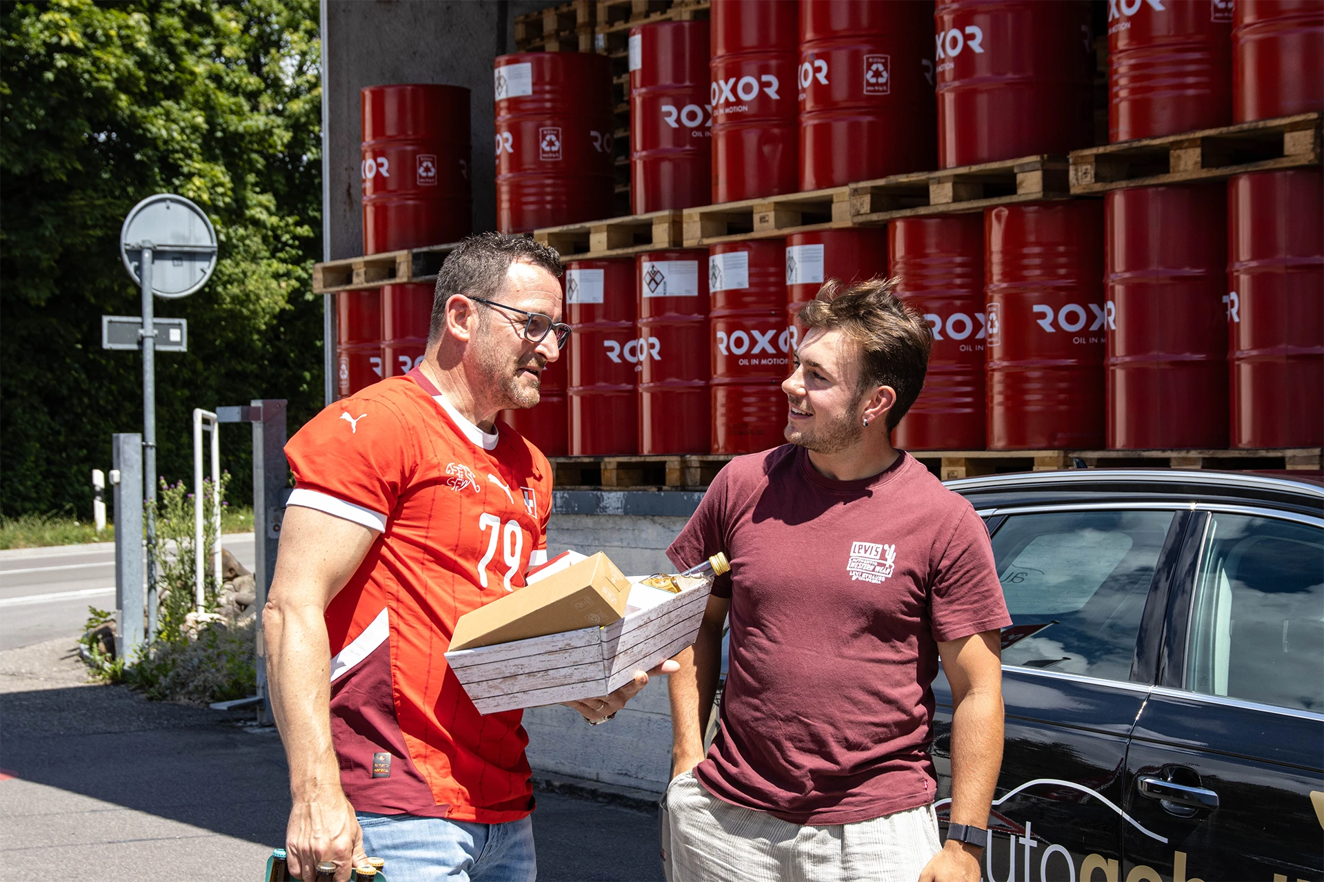 Zwei Männer unterhalten sich lächelnd draußen. Der Ältere im roten Trikot Nr. 79 hält eine Holzkiste mit Geschenken, der Jüngere trägt ein weinrotes T-Shirt. Im Hintergrund: rote Fässer „ROXOR OIL IN MOTION“ auf Paletten, teils schwarzes Auto.