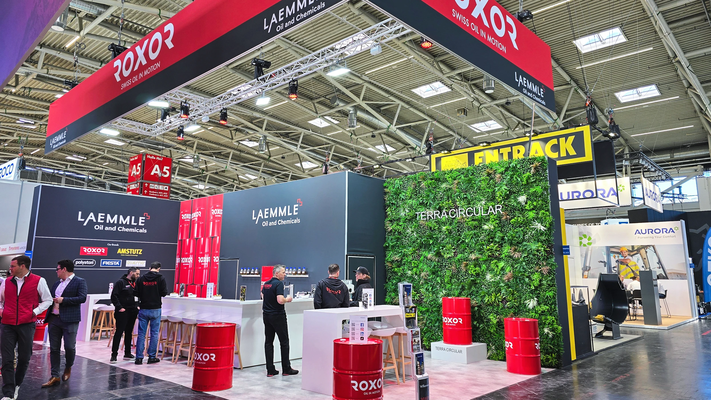 Laemmle Oil and Chemicals’ exhibition stand in a bustling exhibition hall with dark walls and red Roxor barrels. Visitors and stand staff interact at the counters. A green living wall featuring ‘Terracircular’ and a Roxor banner.