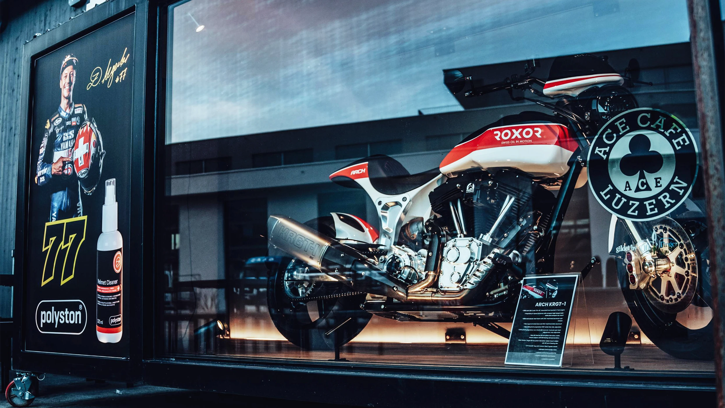 White/red ARCH KRGT-1 custom bike with ROXOR branding in a light glass display case. Left: poster of smiling racing driver in helmet (Swiss cross) and Polyston helmet cleaner. Right: black ACE CAFE LUZERN logo and shiny exhaust.