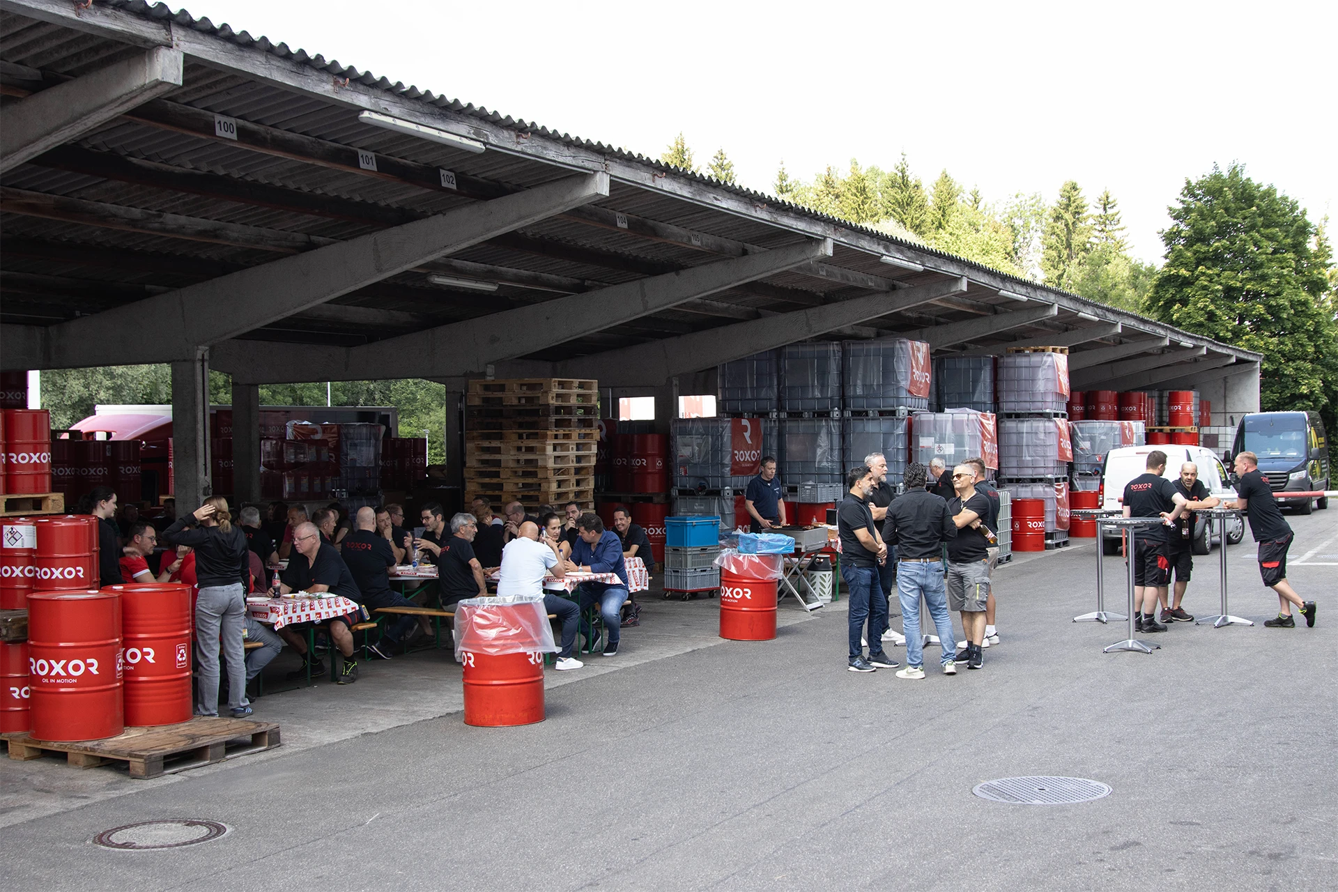 Menschen versammeln sich unter einem großen Wellblechdach im Außenbereich. Einige sitzen an Tischen mit Essen und Getränken, andere stehen und reden. Rote ROXOR-Fässer und gestapelte graue Industrietanks deuten auf ein Firmenevent in einer Lagerhalle.
