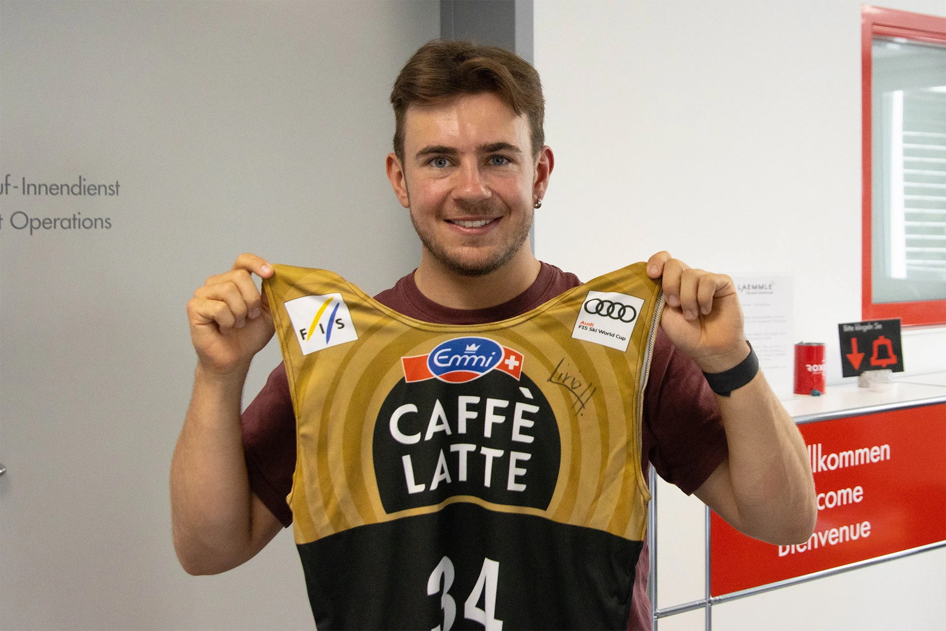 Ein junger Mann mit Bart und rotem T-Shirt lächelt und hält ein gold-schwarzes Skirenntrikot mit den Logos "CAFFÈ LATTE", Emmi, FIS und Audi FIS Ski World Cup sowie der Nummer 34 hoch. Im Hintergrund ist ein Empfangsbereich.