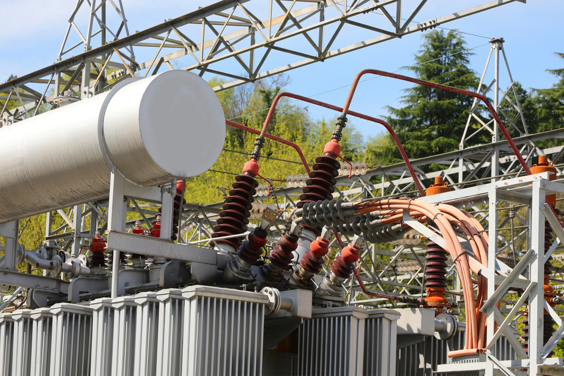 Nahaufnahme eines großen Stromtransformators im Umspannwerk: graue, geriffelte Seiten, oben dicke rote und braune Keramikisolatoren mit orangefarbenen Kabeln. Zylindrisches Oberteil, Gittermasten, Bäume, blauer Himmel.