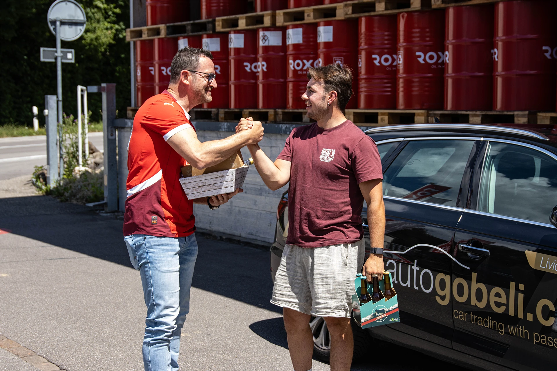 Zwei Männer schütteln sich die Hände und lächeln bei einer Übergabe. Links im rot-weissen T-Shirt hält einer eine kleine Holzkiste, rechts im weinroten T-Shirt trägt der andere einen Sixpack Bier. Dahinter rote Ölfässer und ein Auto.