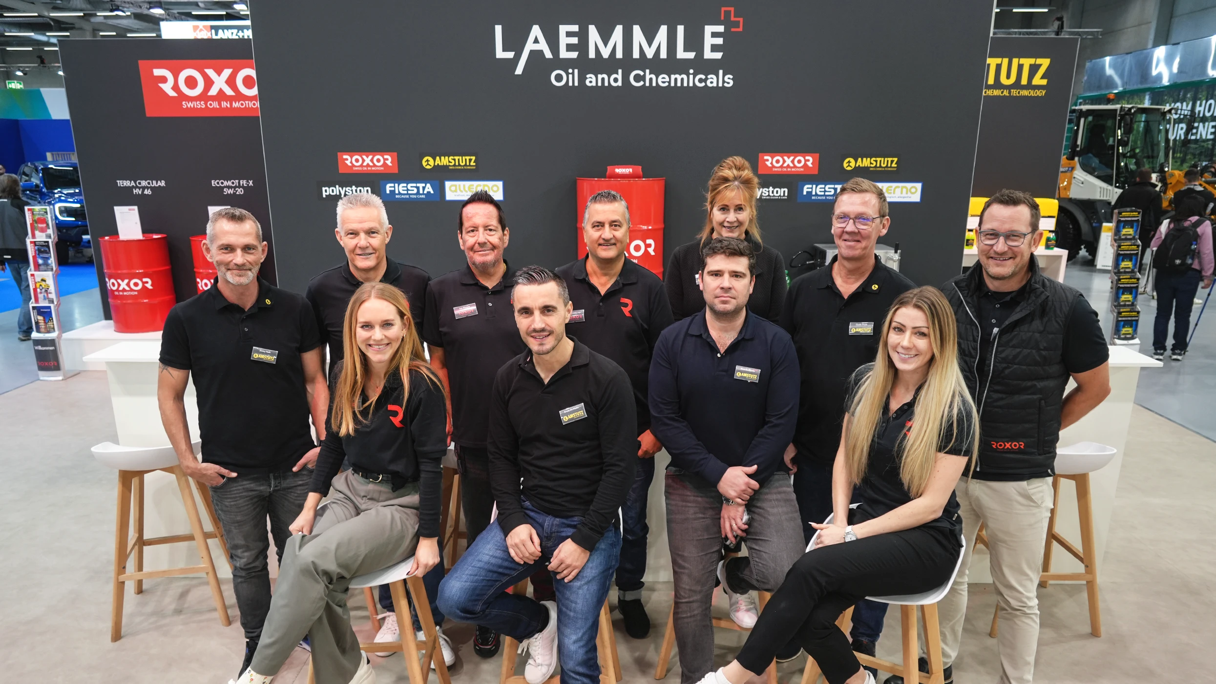 Twelve smiling men and women, most of them wearing black polo shirts, are posing in front of a dark grey exhibition stand. In the background, the logos of Laemmle Oil and Chemicals, Roxor and Amstutz can be seen, along with red oil drums and shelves.