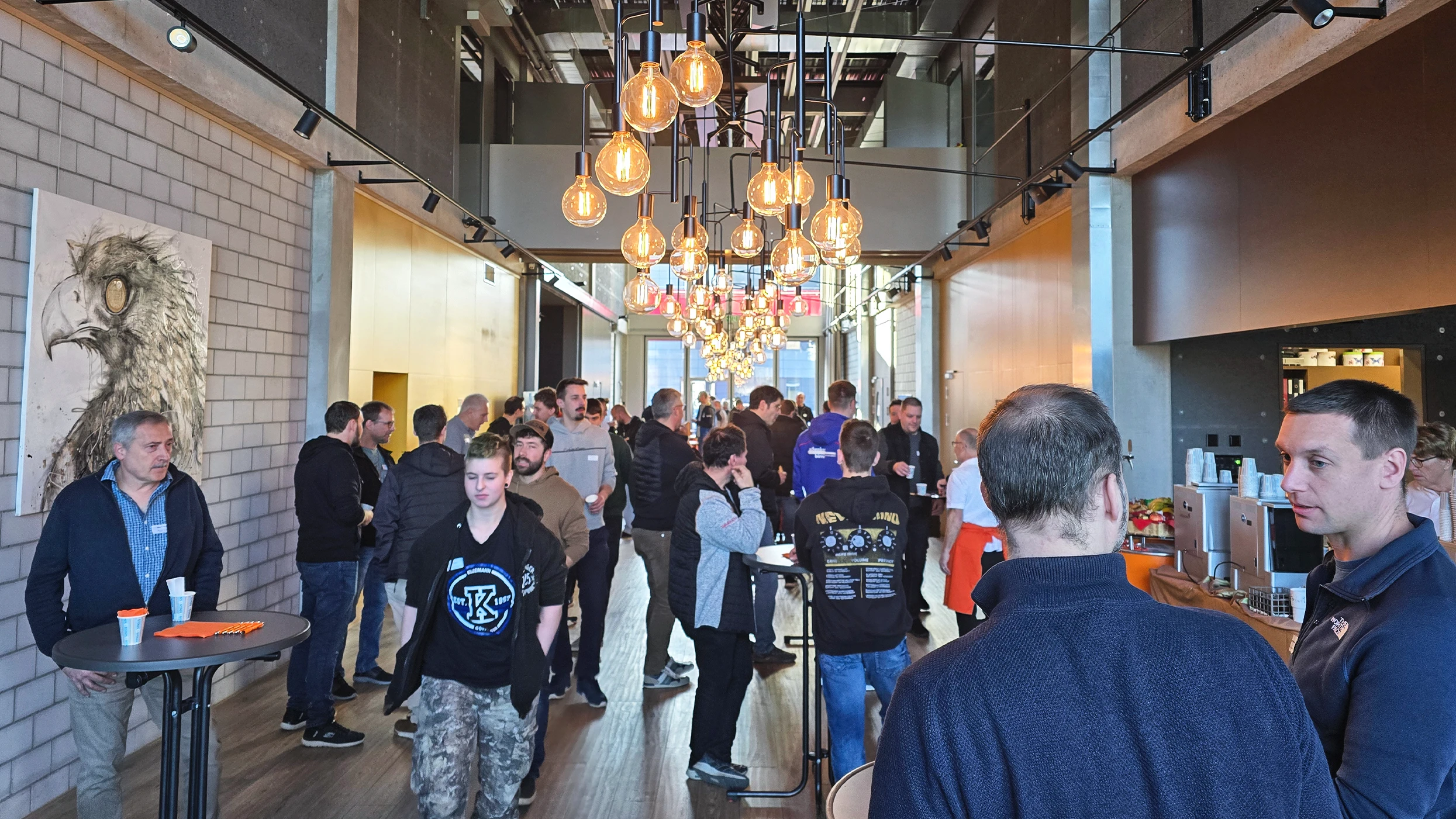 A crowd chats in a modern high-ceilinged room with concrete beams and brick walls. Pendant lights with warm bulbs light the space. Left hangs art of an eagle’s head. Guests chat at bar tables.