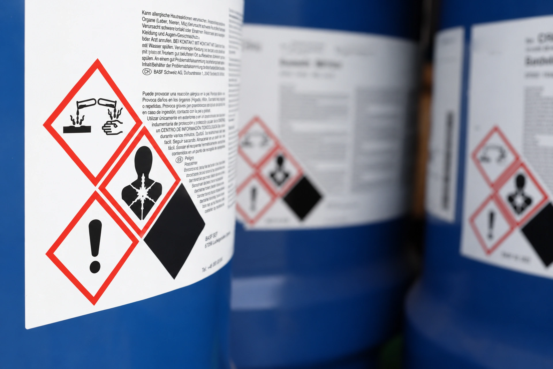 Nahaufnahme eines blauen Industriebehälters mit weißem Gefahrstoffetikett. Drei rote Rauten mit schwarzen Piktogrammen: Ätzwirkung auf Haut/Metall, Gesundheitsgefahr (Organ-/Atemwegsschäden) und Warnung (Ausrufezeichen). Text auch Deutsch/Spanisch.