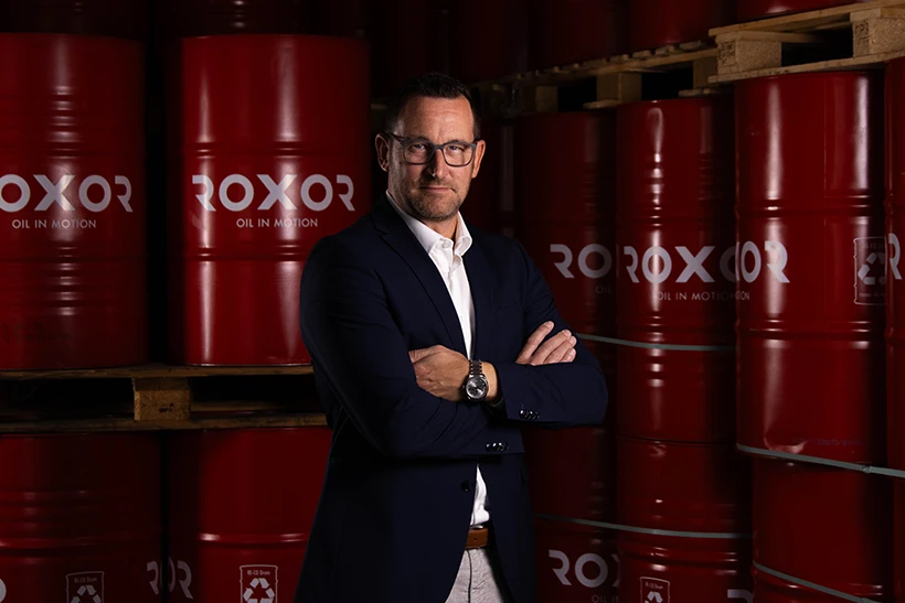 Ein Mann in Anzug und Brille steht mit verschränkten Armen vor einem Lagerraum mit gestapelten roten Fässern. Darauf steht das weiße Logo "ROXOR OIL IN MOTION". Er blickt in die Kamera, wirkt selbstbewusst.