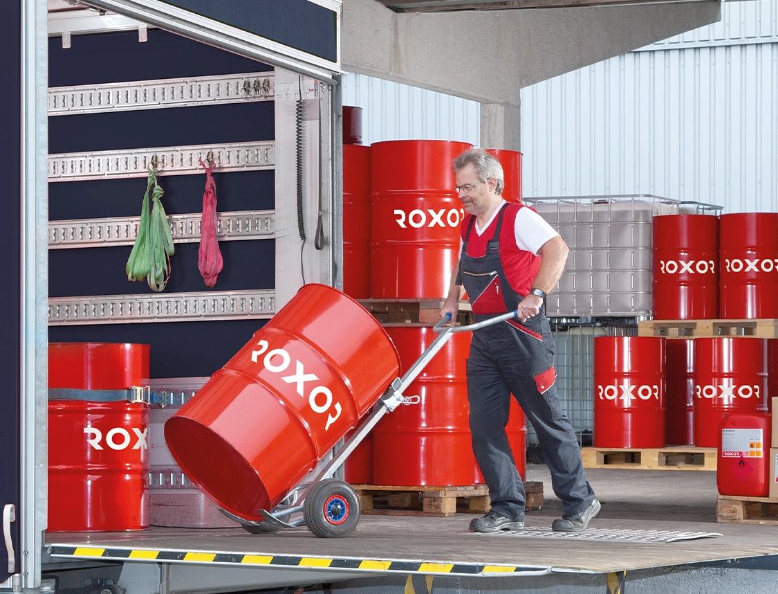 Un ouvrier en tenue de travail pousse un grand fût rouge « ROXOR » sur un diable depuis un quai de chargement. À l’arrière-plan, d’autres fûts rouges sont posés sur des palettes, à côté d’un conteneur IBC argenté dans l’entrepôt.
