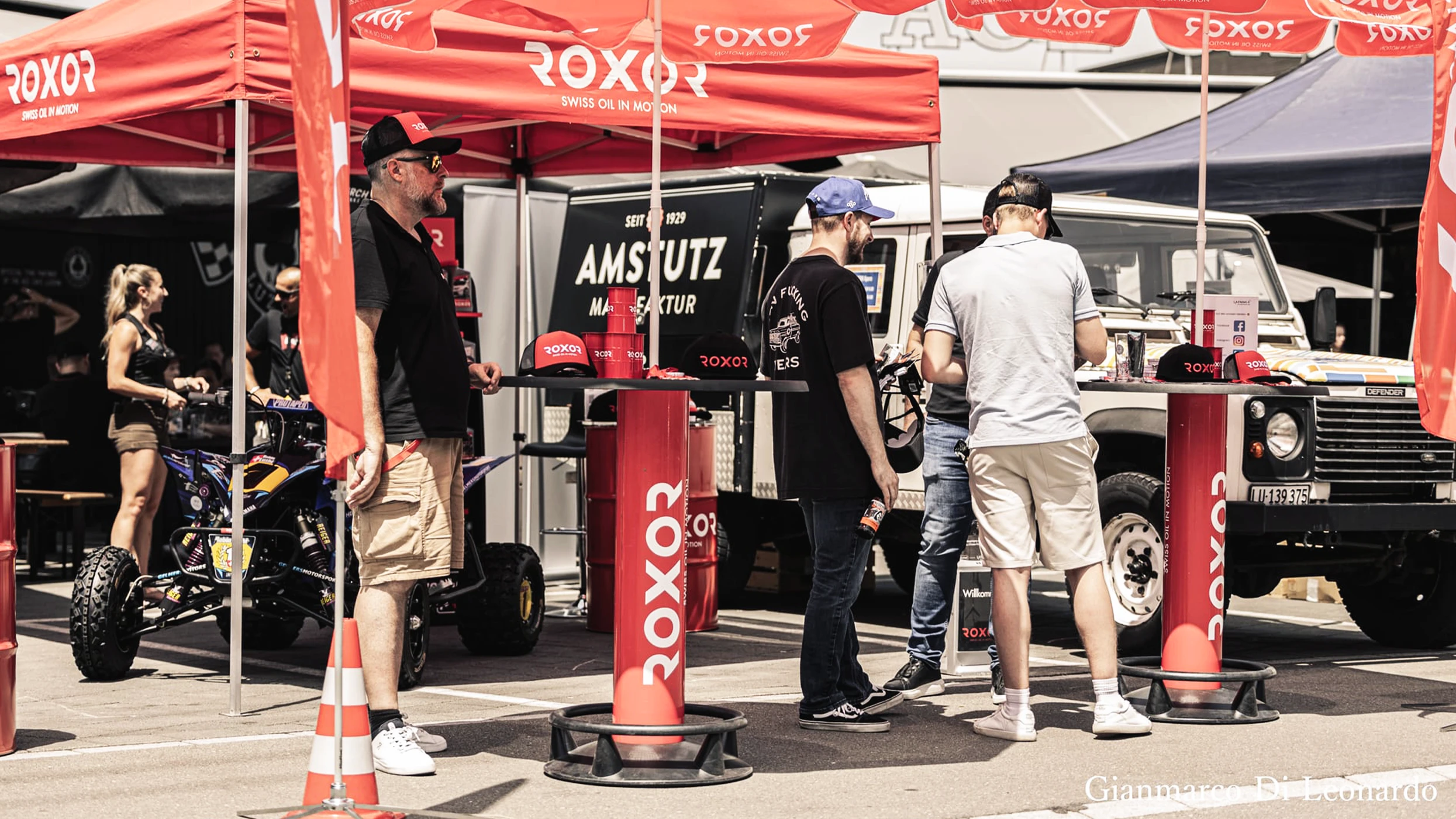 Visitors under red ROXOR pavilions. A man wearing sunglasses in the foreground. People at bar tables next to a quad bike and a white Land Rover Defender. ROXOR flags, the ‘Amstutz Manufaktur’ stand.