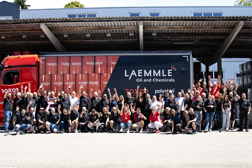 Große Gruppe lachender Mitarbeiter steht und hockt vor rotem Lastwagen mit „LAEMMLE Oil and Chemicals“ auf schwarzem Anhänger. Viele winken. Im Hintergrund Industriegebäude und Laderampe. Rote Fässer zeigen das ROXOR-Logo.