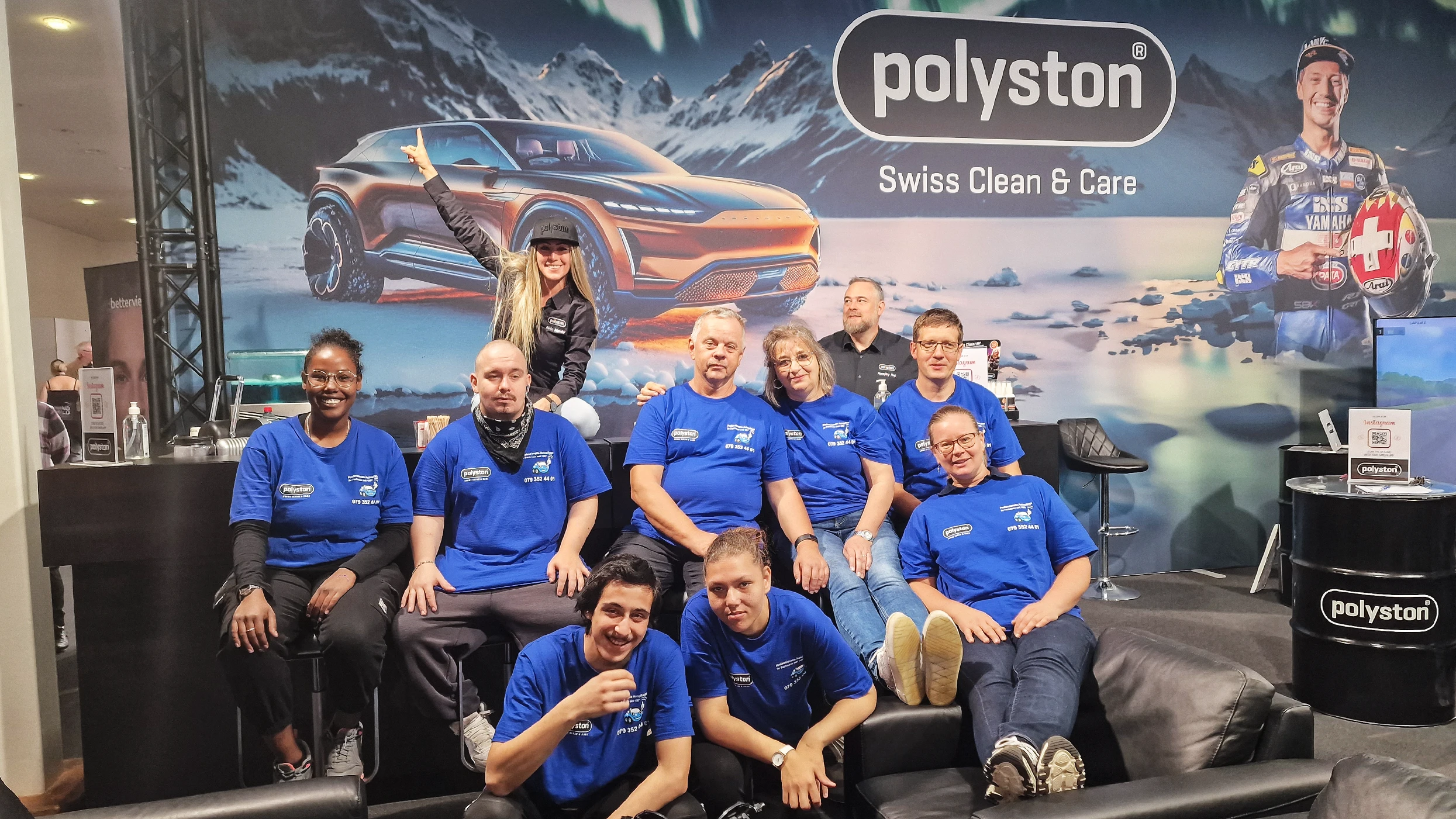 Ten Polyston employees in blue T-shirts pose before a promo backdrop. Some sit on a black sofa while others stand. The backdrop shows a futuristic SUV in a winter scene, the Polyston logo and a racing driver. A woman in black points at the SUV.