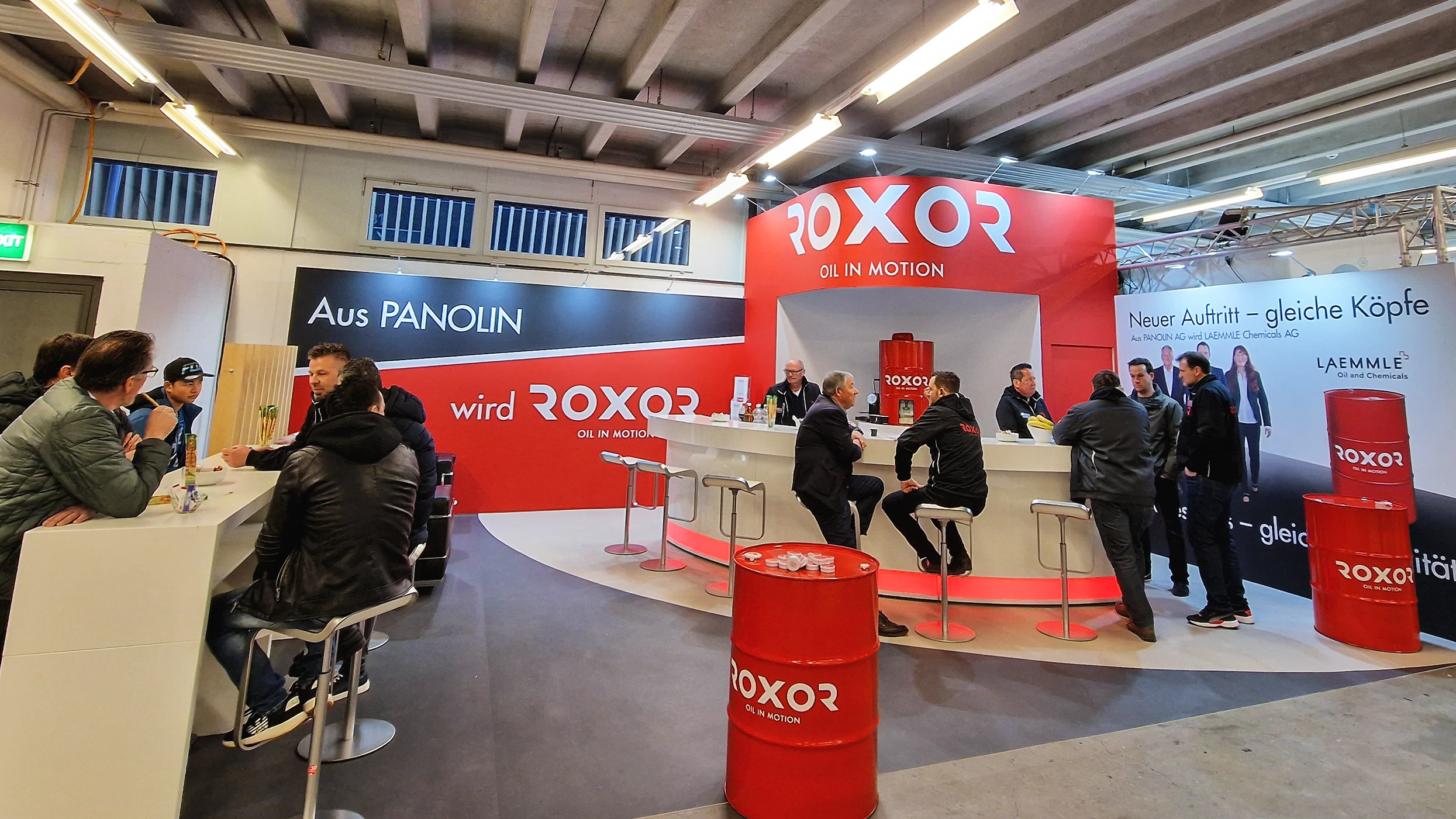 A bustling exhibition stand: people are standing at a curved white bar, chatting. Red walls with ‘ROXOR OIL IN MOTION’ in the background. Red barrels featuring the logo serve as decoration. Business interactions taking place in a brightly lit hall.