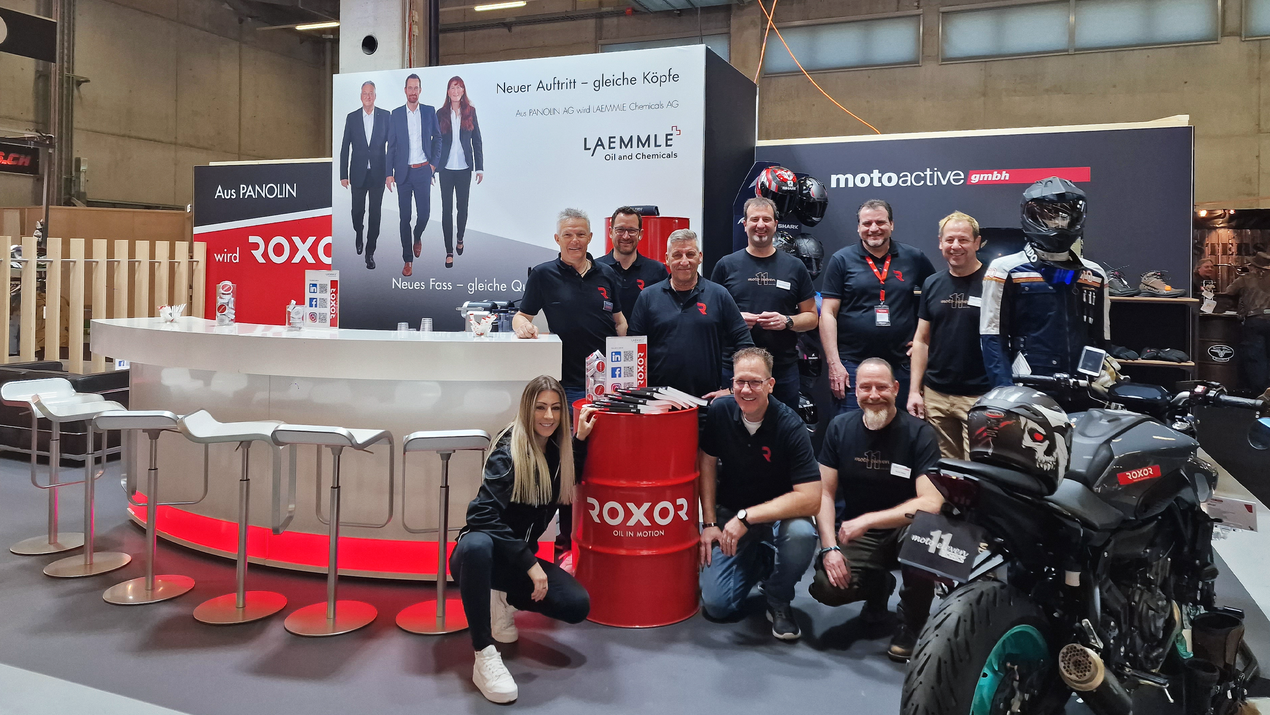 Smiling people at an exhibition stand present the new ROXOR brand. A woman kneels by a red drum marked ‘ROXOR OIL IN MOTION’, while men stand at a white bar and a black motorbike. Banner: Transition PANOLIN to ROXOR/LAEMMLE.