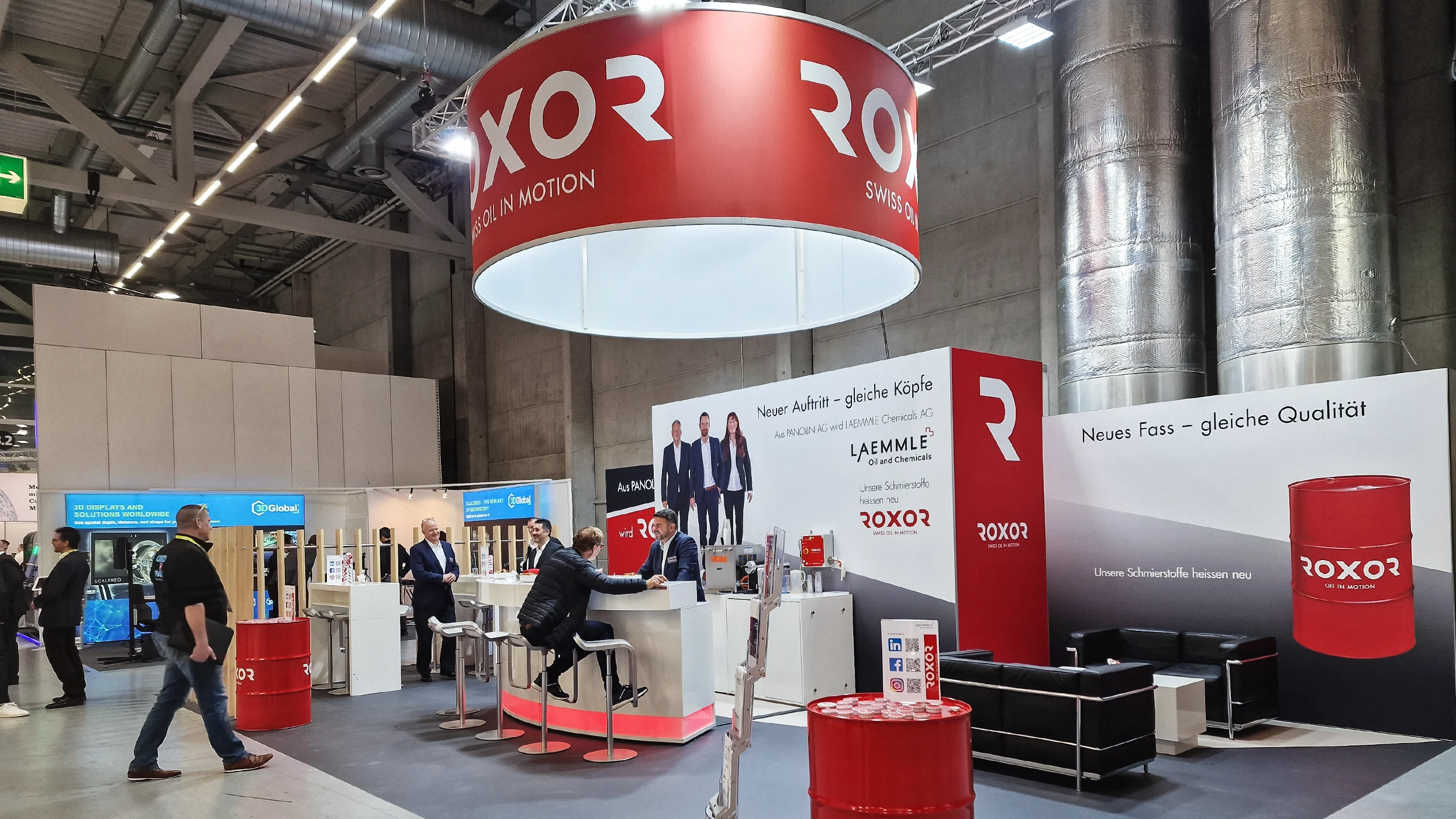 A bustling Roxor exhibition stand in a large hall, featuring a red ceiling banner and information boards. Visitors are chatting at a counter; on the walls are new branding and products such as lubricants in red drums. Ceiling structures are visible.