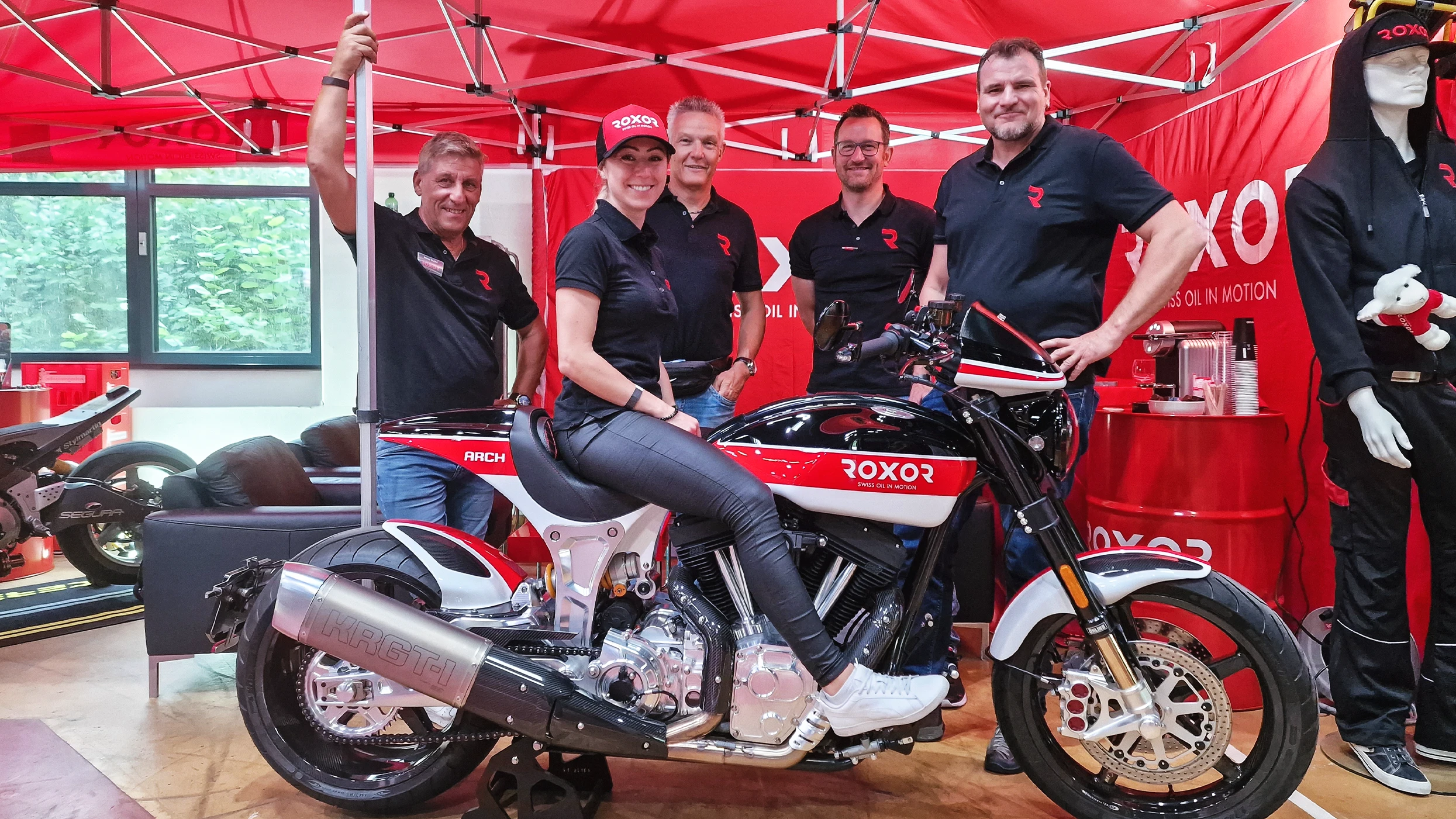 Five smiling people, including a woman on a black ROXOR motorbike with red and white accents, pose in front of a red exhibition stand. They wear black branded clothing with the R logo. A ROXOR banner reads ‘SWISS OIL IN MOTION’.