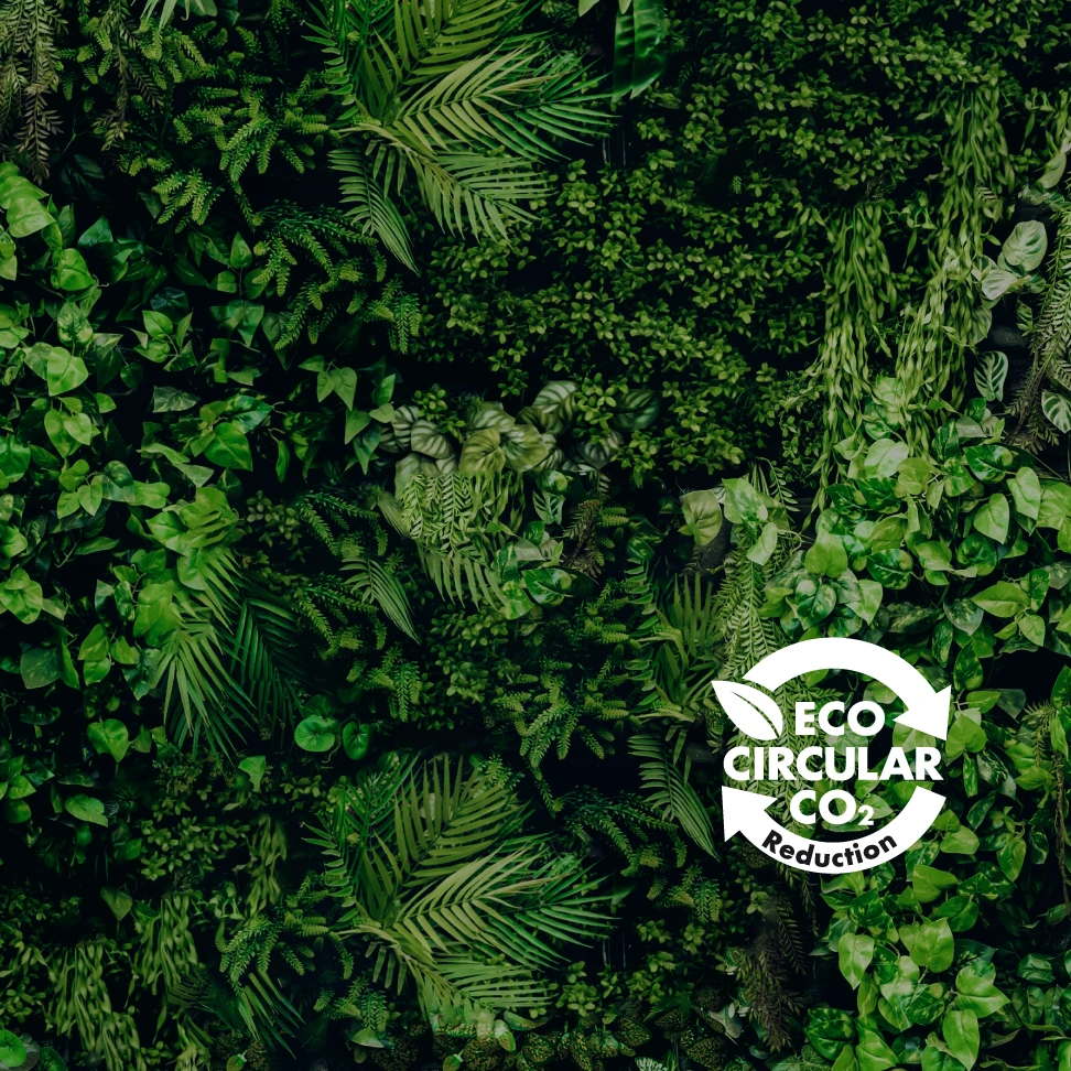 Eine Wand aus grünen Kunstpflanzen wie Farne, Efeu und kleine Blätter bildet ein dichtes, natürliches Muster. Unten rechts ist ein weißes Logo mit kreisförmigen Pfeilen, Blatt und dem Text „ECO CIRCULAR CO2 Reduction