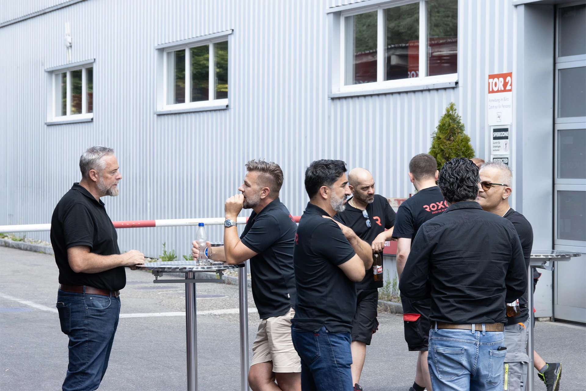 Männergruppe unterhält sich draussen vor hellgrauem Gebäude mit Wellblechfassade und Fenstern. Sie tragen legere Kleidung. Einige stehen um einen Stehtisch mit Getränken. Im Hintergrund: rot-weisse Schranke und Schild „TOR 2“.